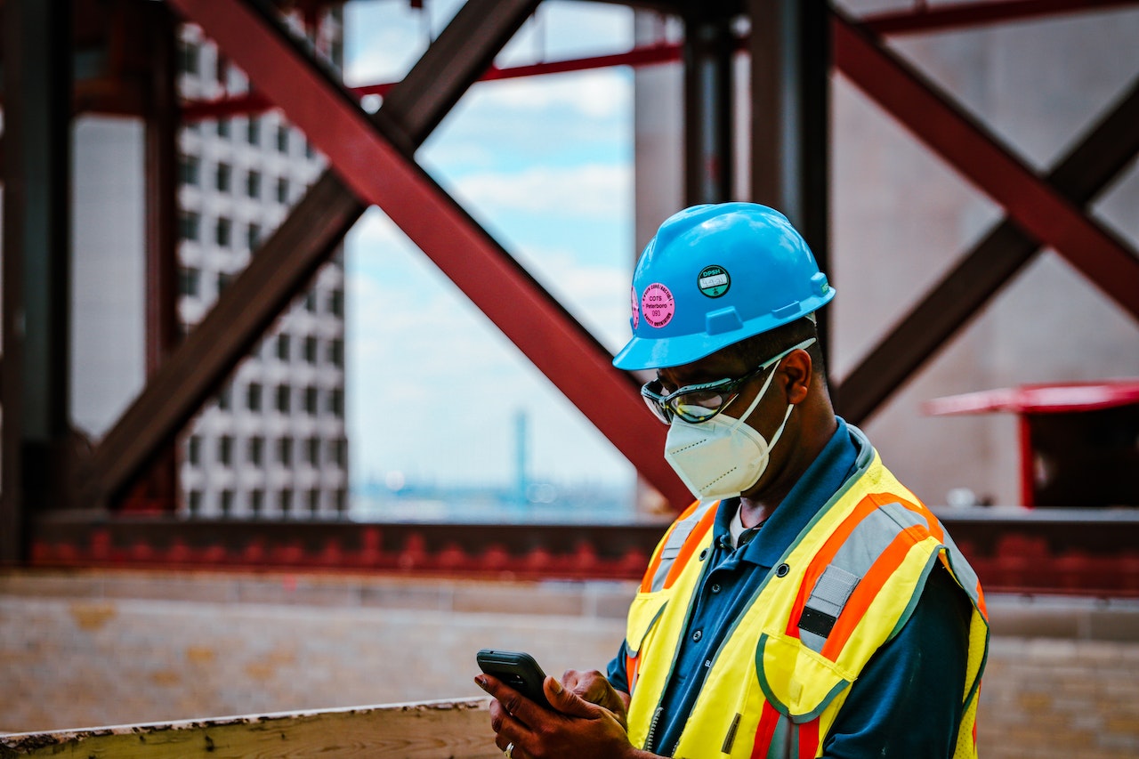 A construction worker in a yellow vest, blue hat, and face mask, checking his phone