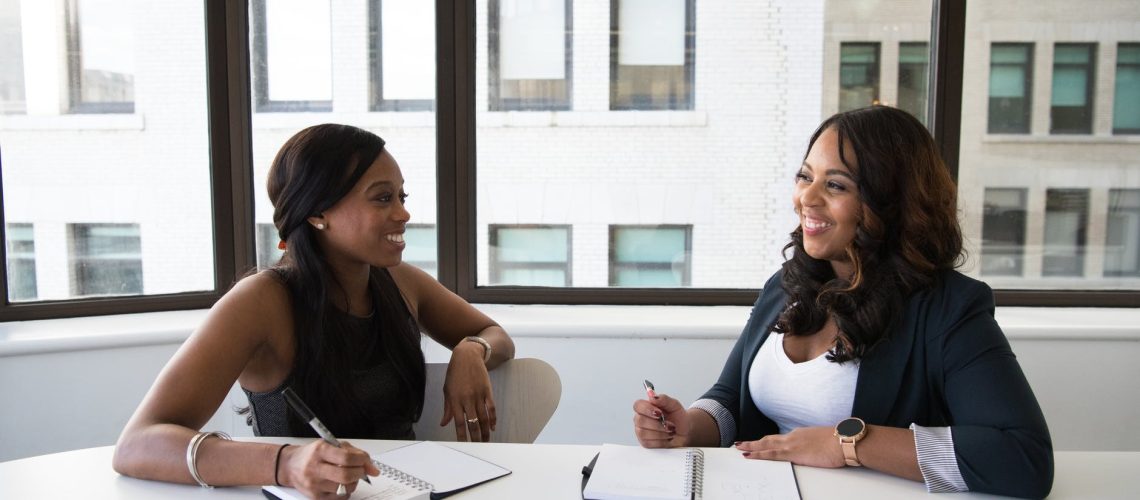 Two women sitting on chairs near a table, smiling at each other