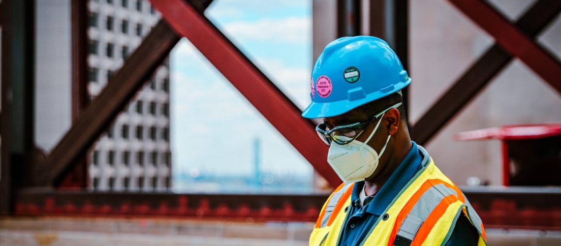 A construction worker in a yellow vest, blue hat, and face mask, checking his phone